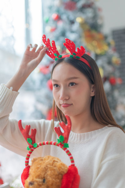 一个年轻美丽友好的亚洲女性女士的肖像照片可爱的圣诞驯鹿发带和柔软的玩具微笑愉快地在一个漂亮的装饰圣诞树的背景前图片下载