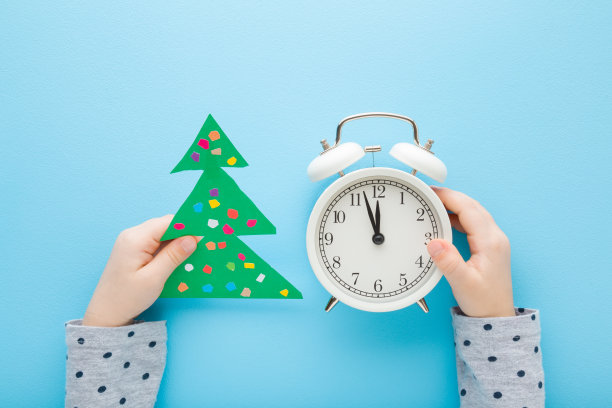 孩子们双手捧着绿色的圣诞树，用彩色的纸和白色的闹钟在蓝色的桌子背景上。柔和的颜色。新年前的午夜倒计时。自顶向下视图。图片下载