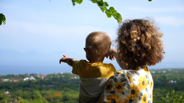 一个怀里抱着孩子的女人的后视图，她站在森林的山顶上，用手指着她的小孩，看远处低地的房子。图片下载