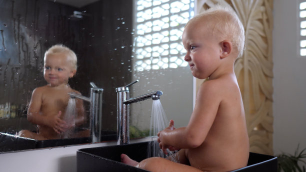 开朗的金发小孩没穿衣服坐在大浴室的水槽里。暖暖的水从水龙头里流出来，宝宝欢快而放肆地把水泼在镜子上。图片下载