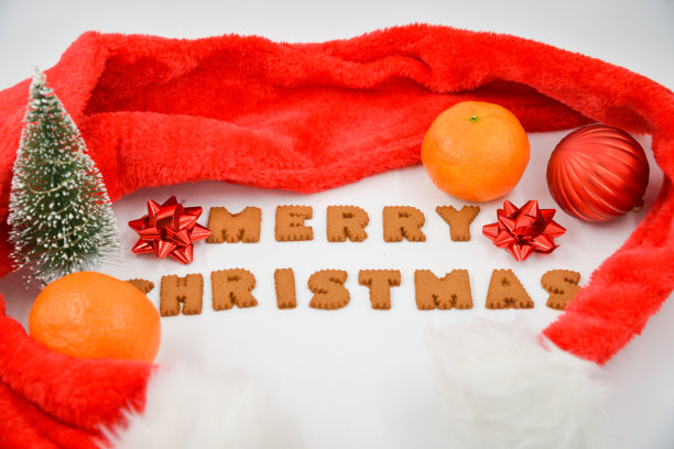 姜饼上写着“圣诞快乐”，白色背景上有一顶红色的圣诞帽，上面有红色的圣诞装饰品和橘子。绿色圣诞树图片下载