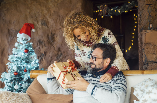 幸福的夫妇享受圣诞节分享礼物在家里休闲活动。圣诞装饰和树的背景。男人从他身后的妻子那里收到一盒惊喜礼物。人们和礼物的圣诞节图片下载