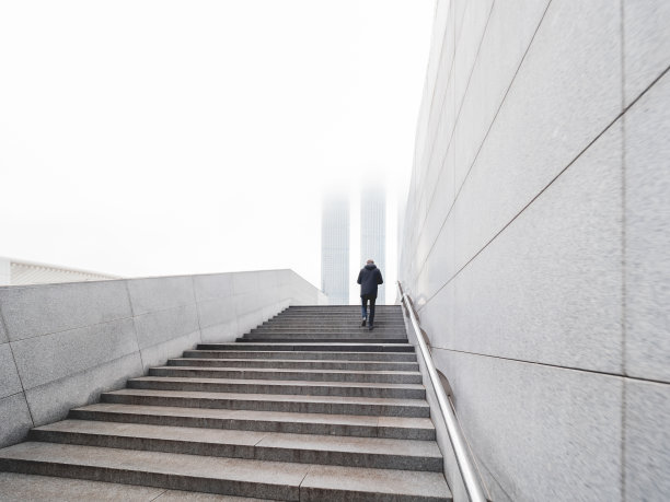 男子在地下人行横道上楼。摩天大楼的顶部笼罩在雾中。大城市里的孤独。城市建筑与孤独的人。单色背景。图片下载
