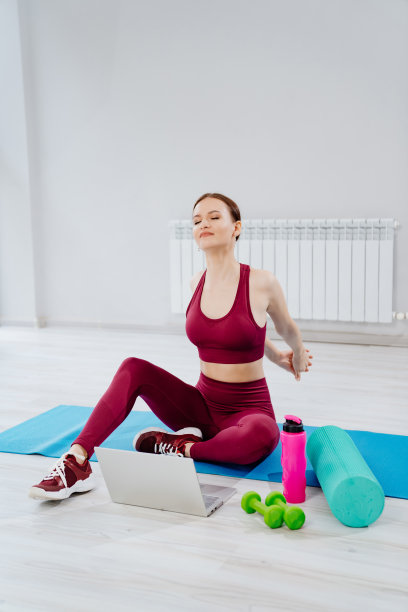一个穿着酒红色运动服的美丽运动女性，笔记本电脑放在健身房的垫子上。图片下载