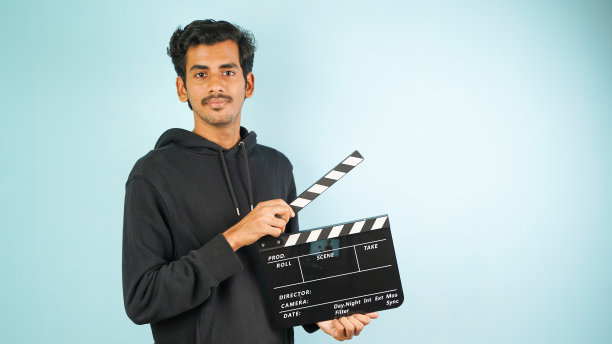 开朗的年轻亚裔印度男子站着拿着隔板，隔板用于电影制作，孤立在彩色背景的工作室肖像图片下载
