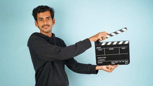 开朗的年轻亚裔印度男子站着拿着隔板，隔板用于电影制作，孤立在彩色背景的工作室肖像图片下载