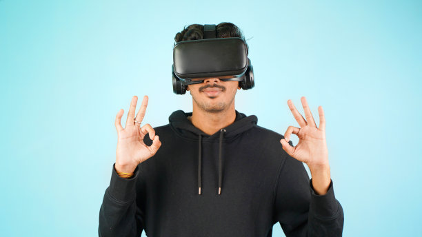 年轻的印度亚裔男子使用VR耳机，眼镜或护目镜，隔离在蓝色室内工作室背景，说好的手势，快乐或幸福图片下载