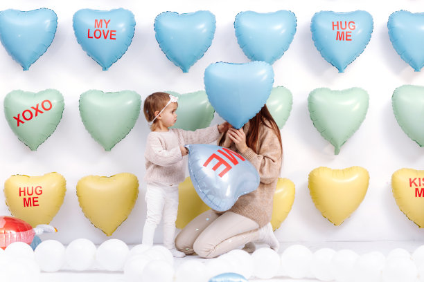 年轻漂亮的妈妈和可爱的小女儿一起站在彩色的心形气球背景上，拥抱和亲吻。生日，情人节，母亲节图片下载