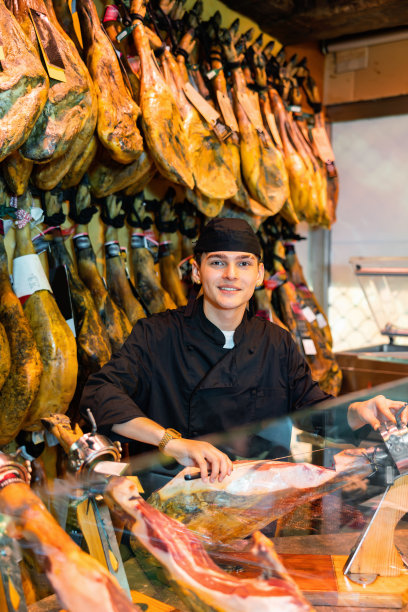 在肉店里，穿着黑色制服的年轻小贩正在切伊比利亚火腿片图片下载