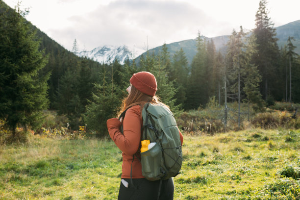 时尚女子穿着绿色背包和红色帽子的背影，在自然中放松时看着山景。旅行和漫游的概念。神奇的寒冷时刻图片下载