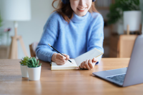 一名十几岁的亚洲女性在图书馆的木桌上使用电脑，戴着耳机，用笔记本电脑通过视频会议在线学习图片下载