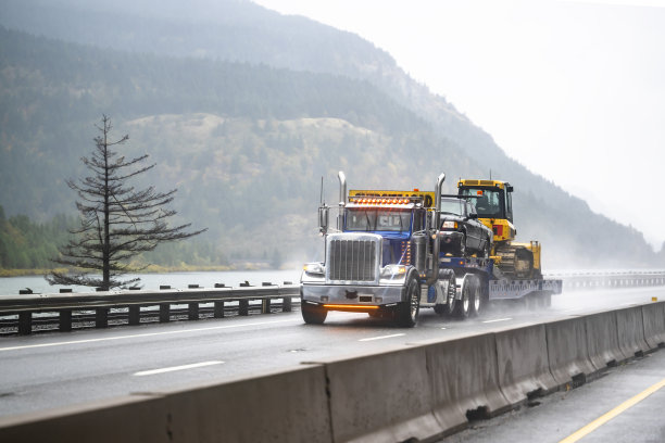 强大的亮蓝色大卡车经典半挂车牵引车，车顶上有超大载重标志，在台阶上运输重物，半挂车在潮湿的道路上行驶，雨水和灰尘图片下载