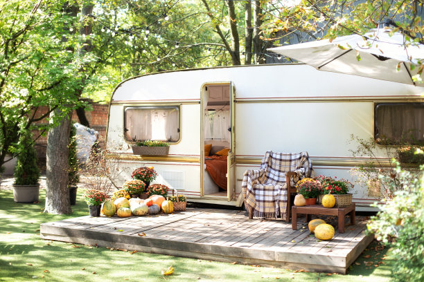 木制房车屋门廊与花园家具。木制扶手椅靠近外面的大篷车拖车。花园露营地。万圣节设计之家。室内舒适的庭院营地与秋季花卉盆栽和南瓜图片下载