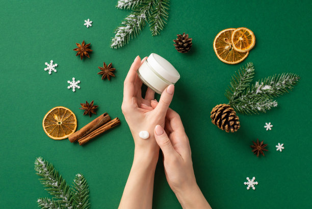 第一人称俯视图照片，女子拿着罐子，把奶油涂在她的手上松果干橘子片肉桂棒八角雪花冷杉树枝在霜孤立的绿色背景图片下载
