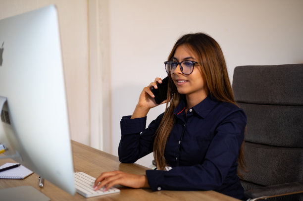 戴眼镜的女商人用她的手机打电话，看着她的电脑屏幕。图片下载