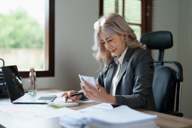 这是一名年轻的亚洲女性的照片，她在凌晨使用她的手机、电脑和桌上的财务文件时露出了微笑图片下载