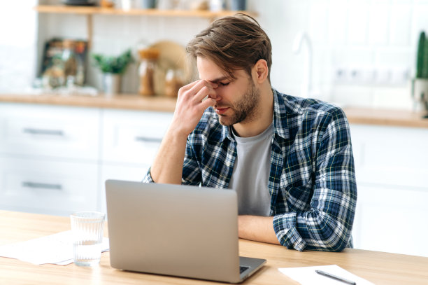 沮丧悲伤的白人男子，闭上眼睛，按摩他的鼻梁，坐在他的工作场所在家里办公室的厨房里，休息。长时间在笔记本电脑前工作，感觉疲惫不堪图片下载