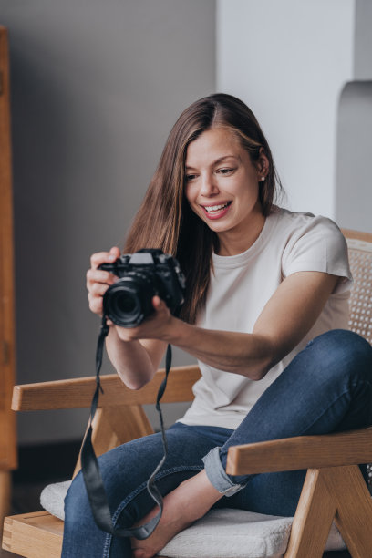 身穿白色t恤和牛仔裤的年轻女子拿着相机看着屏幕，对拍摄结果感到满意。女摄影师坐在椅子上检查相机屏幕上的照片。图片下载