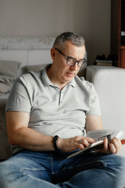 垂直严肃的白发男子戴着眼镜看书，在课本上工作，坐在室内的沙发上。家庭教育图片下载