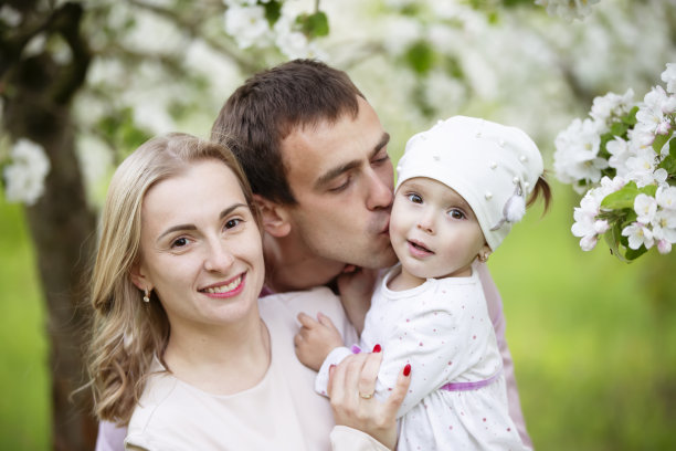 快乐的父母，爸爸妈妈，女儿，年轻的家庭在春天的户外，以开花的树木为背景。图片下载