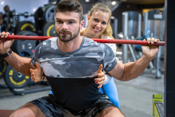 肌肉英俊的男子在深蹲的位置举重与年轻有吸引力的健身女性在健身房的协助。图片下载