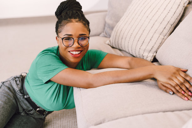 年轻的非裔美国妇女穿着t恤坐在客厅里摆姿势，手在沙发上的股票照片图片下载