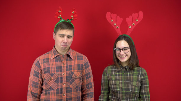一个年轻的女人嘲笑一个年轻的男人，因为他有小角，这个男人被冒犯了，哭了红背景。人们戴着驯鹿鹿角形状的发带。图片下载