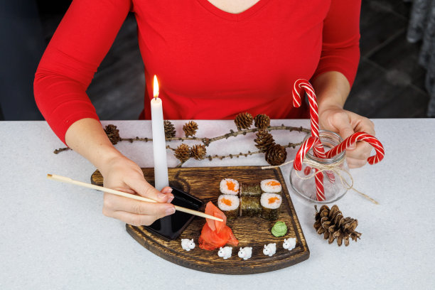 木制砧板上的日式红鱼卷。一个穿红毛衣的女孩用筷子吃寿司。桌上装饰有蜡烛和糖果。圣诞节的概念图片下载