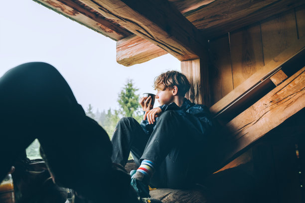 年轻男孩坐在森林木屋的露台上喝着热茶，欣赏早晨云雾缭绕的森林图片下载