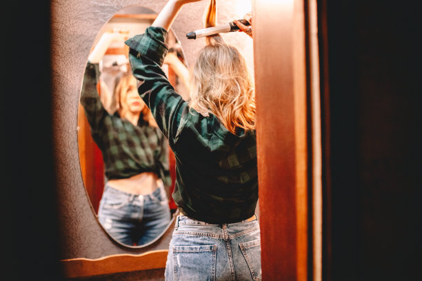 年轻女子用卷发棒看着镜子在家里准备出门图片下载