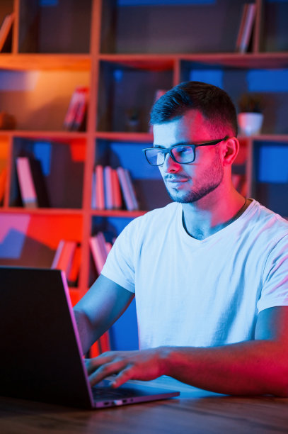 专注于工作。在霓虹灯闪烁的黑暗房间里，戴眼镜、穿白衬衫的男人坐在笔记本电脑旁图片下载