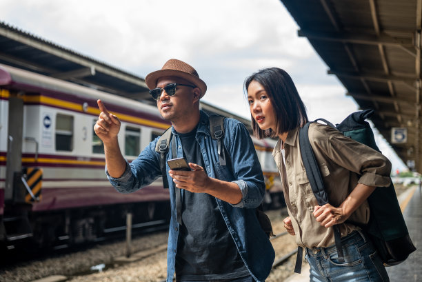 快乐的亚洲帅哥和美女背包客，戴着墨镜，帽子和背包，当火车在火车站迎面而来时，他们指着离开。图片下载