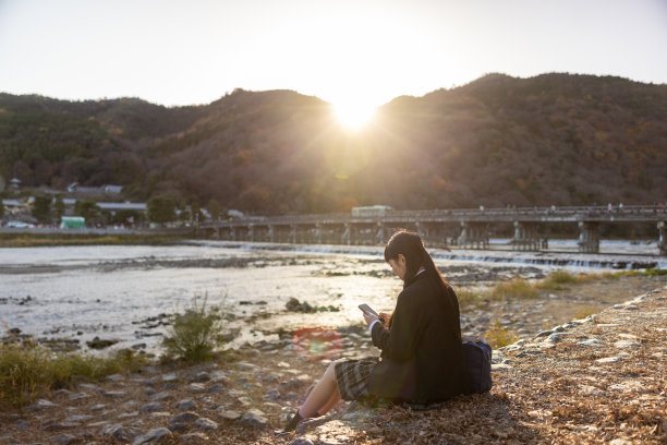高中女孩放学后，日落时分在河边用智能手机图片下载