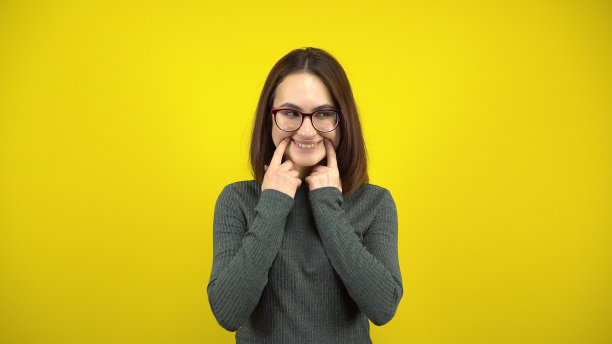 一名年轻女子用手指在黄色背景上露出愉快的微笑。戴眼镜，穿绿色毛衣的女人。图片下载