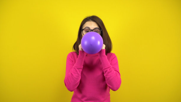 一名年轻女子在黄色背景上用嘴给紫色气球充气。穿粉色高领毛衣戴眼镜的女孩。图片下载
