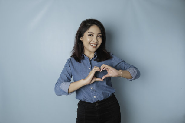 穿着蓝色衬衫的年轻迷人的亚洲女性感到快乐，浪漫的心形手势表达了温柔的感情图片下载
