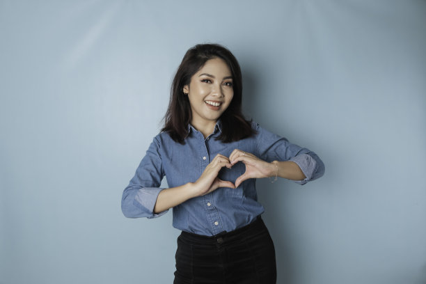 穿着蓝色衬衫的年轻迷人的亚洲女性感到快乐，浪漫的心形手势表达了温柔的感情图片下载