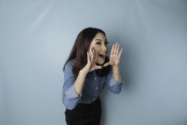 年轻漂亮的女人穿着一件蓝色的衬衫大声喊叫和尖叫一边用一只手在她的嘴。通信的概念。图片下载