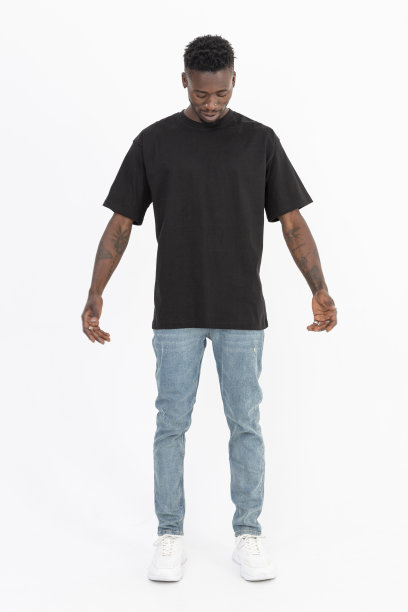 一名身穿深灰色t恤和蓝色牛仔裤的非洲裔美国黑人站在演播室的白色背景前，看着他的t恤。图片下载