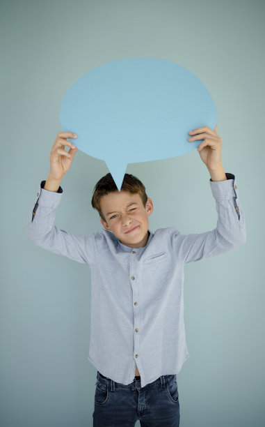 穿着蓝色衬衫和牛仔裤的小男孩站在蓝色背景前，拿着蓝色的语音泡泡图片下载