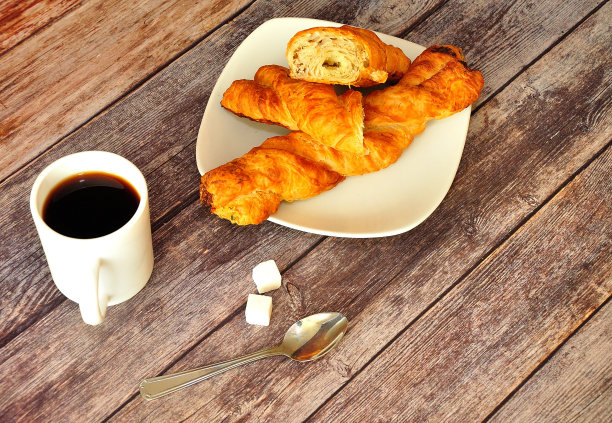 一个装着两个螺旋形酥皮包的盘子，一杯热咖啡，旁边有一把勺子，木桌上放着两块方糖。图片下载