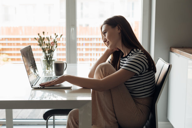 无聊，冷静的年轻棕色头发的女人坐在厨房弯曲的腿，在笔记本电脑上工作，喝咖啡靠窗图片下载