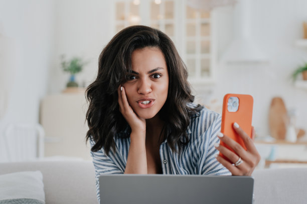 愤怒的非洲裔美国女孩坐在桌子旁，拿着笔记本电脑，拿着手机，看着相机，用震惊的表情触摸脸。迷人的巴西年轻女子惊讶于新闻，销售，折扣。图片下载