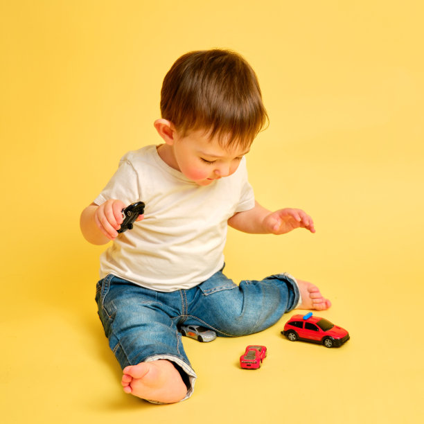 蹒跚学步的婴儿在工作室的黄色背景上玩玩具车。穿着白色t恤和蓝色牛仔裤的快乐孩子在玩玩具车。一岁零四个月的孩子图片下载