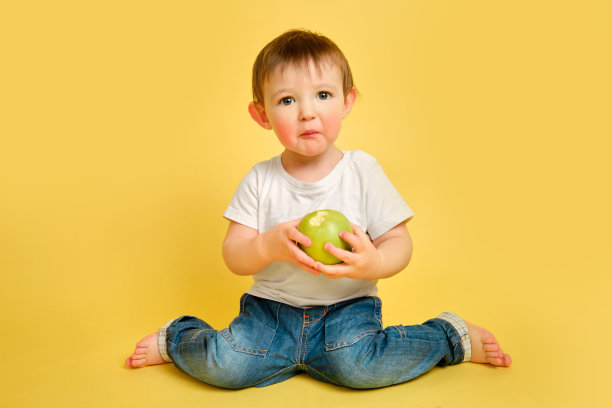 蹒跚学步的婴儿在工作室黄色的背景下吃酸苹果，惊讶的脸。一个穿着白t恤和蓝色牛仔裤的酸溜溜的孩子。一岁零四个月的孩子图片下载