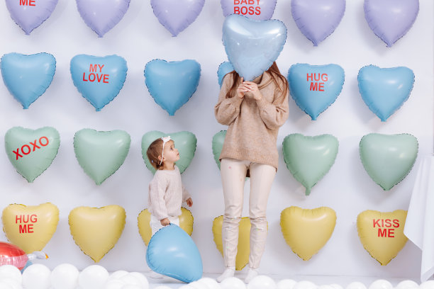 年轻漂亮的妈妈和可爱的小女儿站在彩色的心形气球背景上，拿着蓝色的情人节气球一起玩。生日，情人节，母亲节图片下载