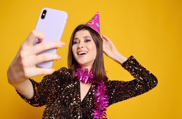 年轻女子戴着粉红色的派对帽，脖子上系着粉红色的蟒蛇，微笑着用智能手机在黄色背景上自拍图片下载
