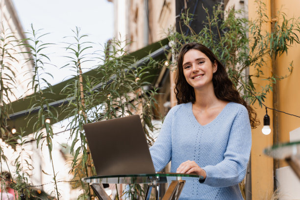 年轻女子在餐厅的笔记本电脑上远程工作。带着笔记本电脑的迷人女孩坐在室外的咖啡馆里，与朋友和同事聊天。图片下载