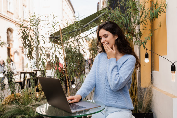 震惊的女孩在网上赌场赢了赌注，坐在户外咖啡馆的笔记本电脑。快乐的惊讶的女人微笑着，看着在网上赌场的收入和欣喜的数额。图片下载