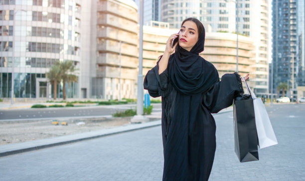 表情严肃的阿拉伯妇女穿着传统服装，手提购物袋在城市街道上边走边打电话图片下载